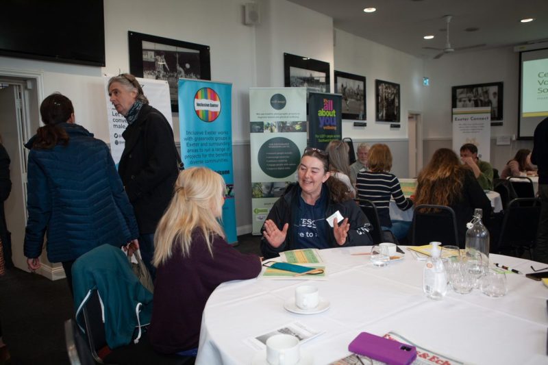 Two women sat chatting at a table, with a few people standing behind them.