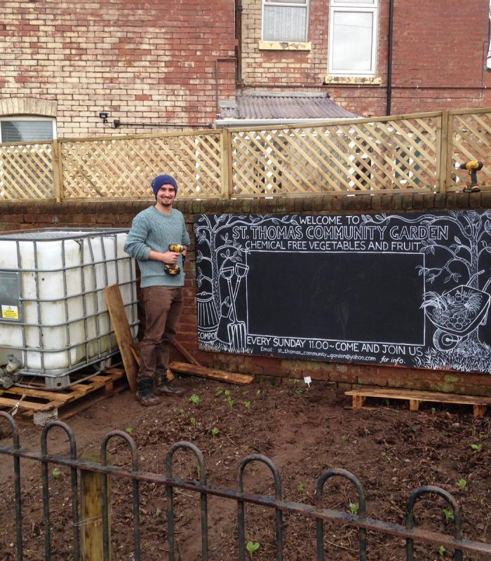 Man standing outside holding a drill next to a chalkboard sign on the wall which says 'Welcome to St Thomas Community Garden. Chemical free vegetables and fruit. Every Sunday 11:00. Come and join us.'